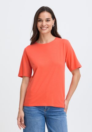 Donna sorridente con lunghi capelli scuri che indossa una maglietta arancione a maniche corte e jeans blu, in piedi davanti a uno sfondo chiaro uniforme.