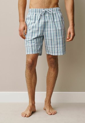 Vyras, vilkintis šviesiai mėlynos, baltos ir žalios spalvos languotus šortus iki kelių su raišteliu, basas stovintis prie neutralaus sienos.