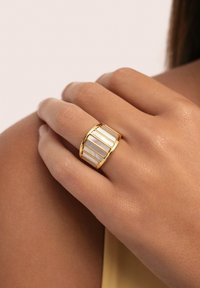 Gouden ring met verticale witte inlegpanelen, met een gestructureerd oppervlak en een gebogen, brede ringontwerp.