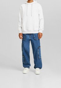 Sudadera con capucha gris con cordones, bolsillo frontal; combinada con vaqueros azules holgados con múltiples parches y cremalleras; zapatillas blancas.