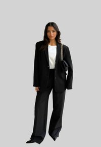 Črn oversize blazer z belo srajco, v kombinaciji z ohlapnimi črnimi hlačami in špicastimi črnimi petkami. Dodatki vključujejo majhno črno torbico.