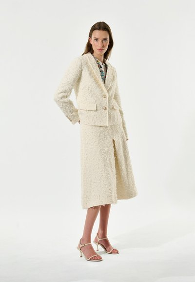 Costume blanc texturé avec une veste ajustée et une jupe au genou, dotée de poches avant et de deux boutons, associée à des escarpins à bride de cheville beige.