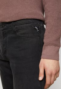 Jeans in denim nero con una vestibilità classica, tasche anteriori e un logo discreto sulla cintura, abbinati a un maglione in mauve a trama.