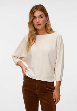 Femme aux longs cheveux blonds portant un haut blanc ample à manches trois-quarts et un pantalon en velours côtelé marron, debout avec une main sur la hanche.