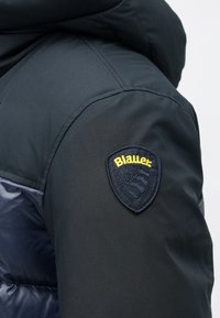 Tissu d'extérieur noir avec une section rembourrée grise, arborant un logo en forme de blason bleu marine et jaune sur l'épaule gauche. Texture lisse.