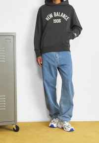 Felpa grigia con cappuccio e scritta "NEW BALANCE 1906", abbinata a jeans larghi azzurri e sneakers bianche con dettagli blu. Sullo sfondo un armadietto metallico.