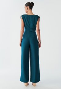 A woman stands facing away, wearing a dark teal jumpsuit with wide legs, styled hair in a bun, and beige high-heeled shoes.