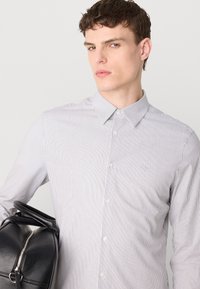 Camisa de botones a rayas en blanco y azul, con cuello, botones frontales y un corte ajustado. El modelo sostiene un bolso de cuero negro.