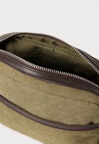 Τσάντα από καμβά σε ελαιοπράσινο χρώμα με φερμουάρ από καφέ δέρμα. Το εσωτερικό είναι επενδεδυμένο με ύφασμα, διαθέτει κλείσιμο με φερμουάρ και εμφανείς λεπτομέρειες ραφής.