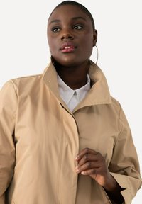 Giacca impermeabile color sabbia con colletto alto e zip frontale. Presenta una texture liscia e una camicia bianca sottostante. Cuciture con dettagli dorati visibili.