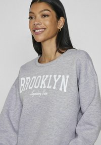 Sudadera gris con "BROOKLYN" en letras blancas en negrita y "Legendary Team" en una fuente más pequeña. El material parece suave y texturizado.