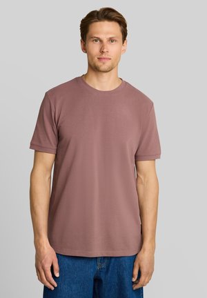 Mann mit kurzen braunen Haaren, der ein mauvefarbenes T-Shirt mit kurzen Ärmeln und blaue Jeans trägt, steht vor einem schlichten hellgrauen Hintergrund.