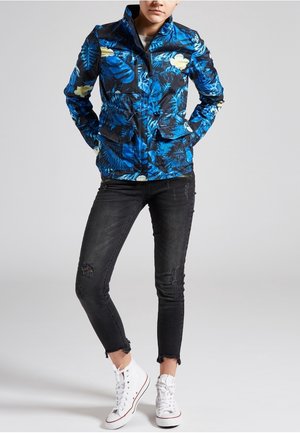 Chaqueta azul con estampado floral, de manga larga y cierre de cremallera, combinada con jeans negros desgastados y zapatillas blancas altas.