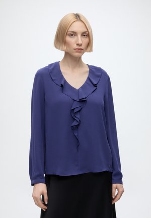 Jeune femme aux cheveux blonds courts portant un chemisier violet à manches longues avec un col en V à volants et une jupe noire, debout sur fond blanc.