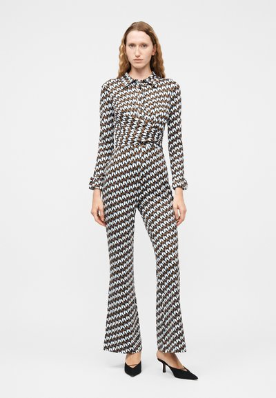 Γυναίκα που φοράει ολόσωμη φόρμα με μακριά μανίκια και γιακά, με γεωμετρικό μοτίβο σε καφέ, λευκό και ανοιχτό μπλε χρώμα, και μαύρες μυτερές γόβες.