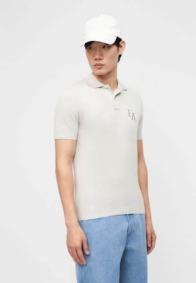 Άνδρας που φοράει ανοιχτό γκρι πόλο μπλουζάκι EA, λευκό καπέλο και ανοιχτό μπλε τζιν, κοιτάζοντας προς τα αριστερά του, με φόντο μια απλή επιφάνεια.