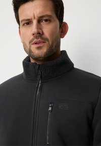 Zwarte jas met rits en fleece voering, voorzien van een hoge kraag, een ritszak aan de voorkant en subtiele logodetails op de linkerborst. Zachte textuur en moderne pasvorm.