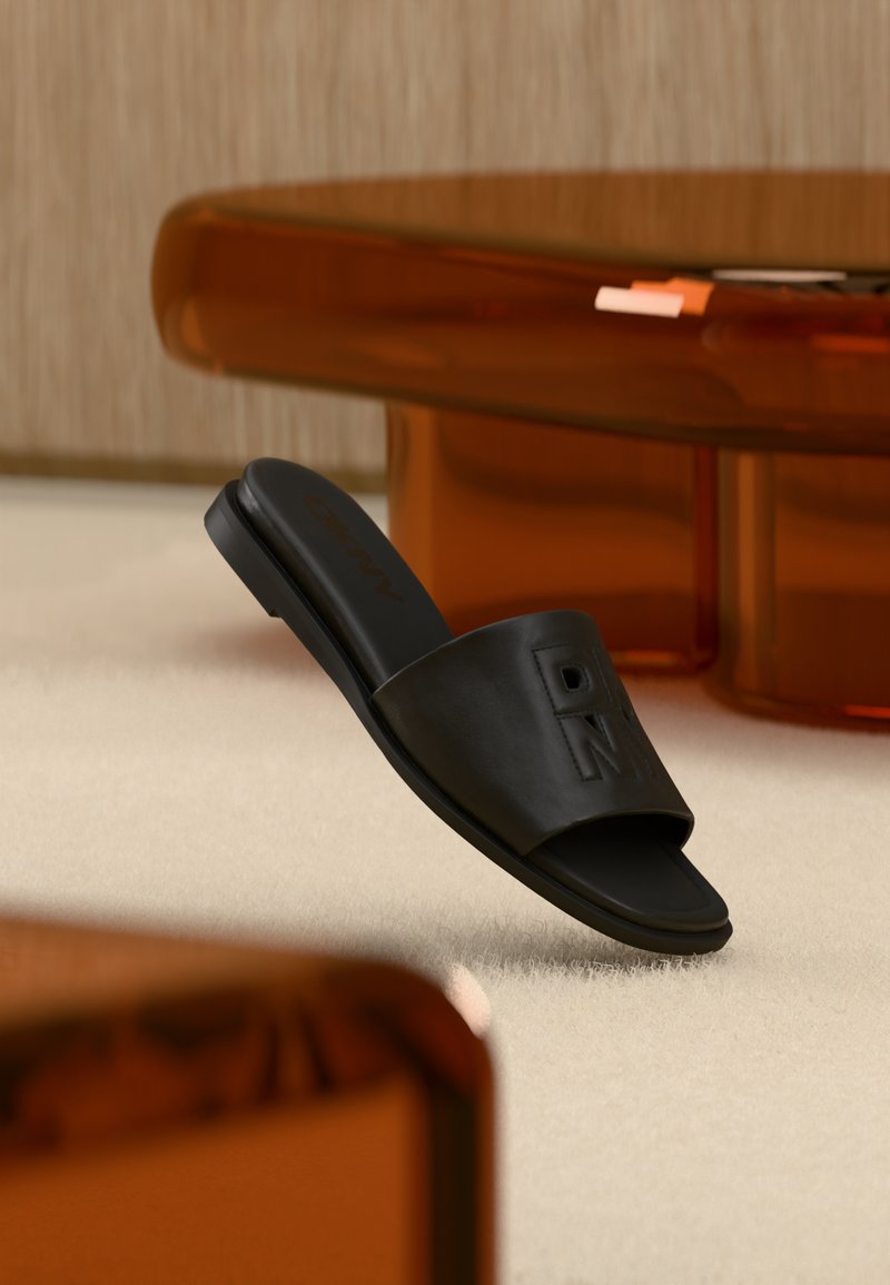 Sandale slide noire avec les lettres embossées "D" et "N" sur la lanière, posée sur un tapis clair près d'une table translucide ambrée.