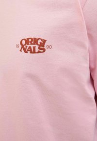 Roosa puuvillane sviiter, millel on rindkerel väike pruun "ORIGINALS 1990" logo. Kanga tekstuur tundub pehme ja sile.