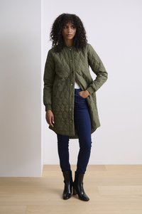 Vadderad, olivgrön lång kappa med tryckknappar och ribbade ärmslut, kombinerad med mörkblå skinny jeans och svarta klackboots.