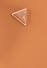 Tan gekorreld leer met een driehoekige goudaccent logo met de tekst "GUESS USA" en een sleutelgat symbool. Soepel, verfijnd ontwerp.