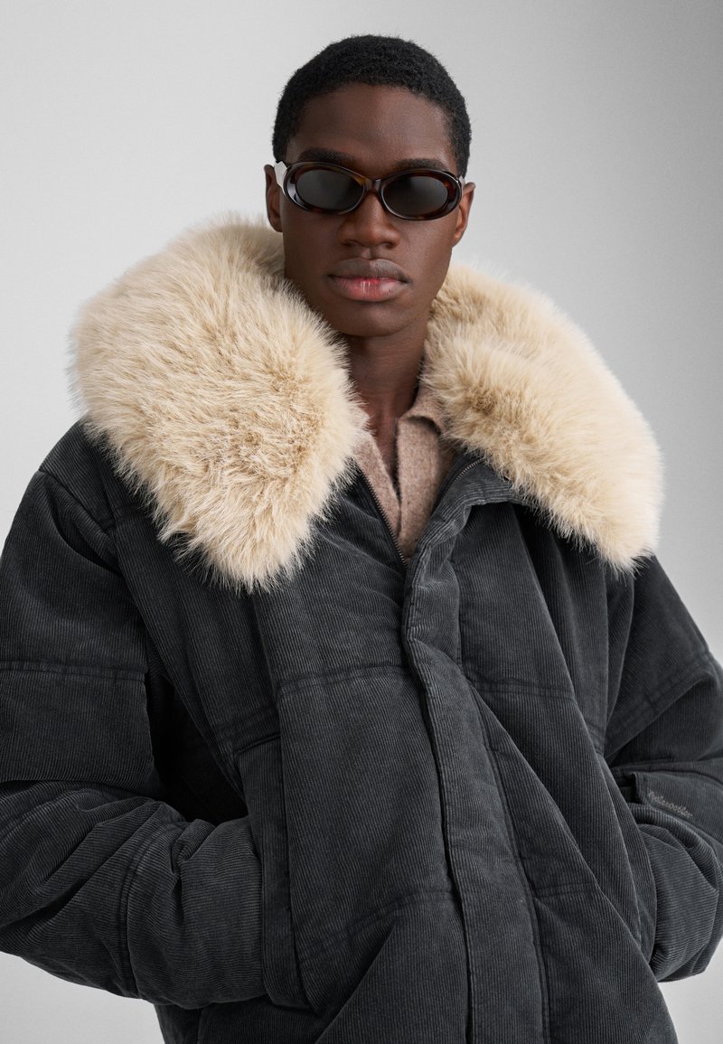 Mørkegrå kordfløjljakke med en stor, fluffy krave i cremefarvet faux-fur. Lynlås foran, oversized pasform og ribbet tekstur.