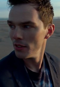 Gros plan sur un homme avec des cheveux courts et ébouriffés, portant un t-shirt noir avec un motif dégradé bleu et un blazer sombre, tourné légèrement sur le côté.