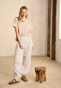 Egy mosolygó nő bézs ingben és csíkos, bő szárú nadrágban áll egy kis, rusztikus fa szék mellett egy semleges tónusú szobában.