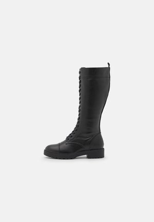 Wysoki czarny skórzany but na sznurowadła z masywną podeszwą i pętelką z tyłu, pokazany z boku na białym tle.