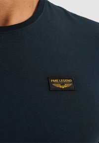 Uski blizak pogled na tamnoplavu majicu s malim crnim i žutim PME Legend pečatom na prsima, koji prikazuje orla i tekst.