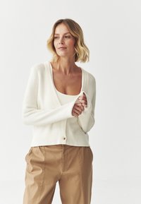 Cardigan bianco a coste con scollatura a V e chiusura a un bottone dorato. Indossato sopra una maglietta di colore chiaro e abbinato a pantaloni beige.
