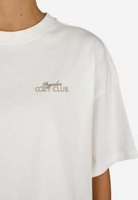 Balta kokvilnas T-kreklus ar nokrišanas kaklu un īsām piedurknēm, ar tekstu "Pegador COZY CLUB" izdrukātu bēšā krāsā kreisajā krūšu daļā.