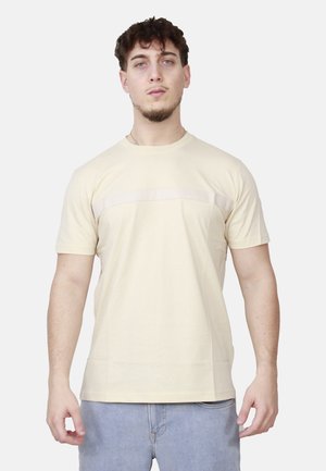 Giovane uomo in piedi che indossa una maglietta a maniche corte color beige chiaro e jeans azzurri chiari su uno sfondo semplice.