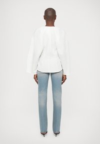 Camisa de manga longa branca com detalhes franzidos nas costas, combinada com jeans retos de perna clara e desbotada e sapatos de salto alto pretos.