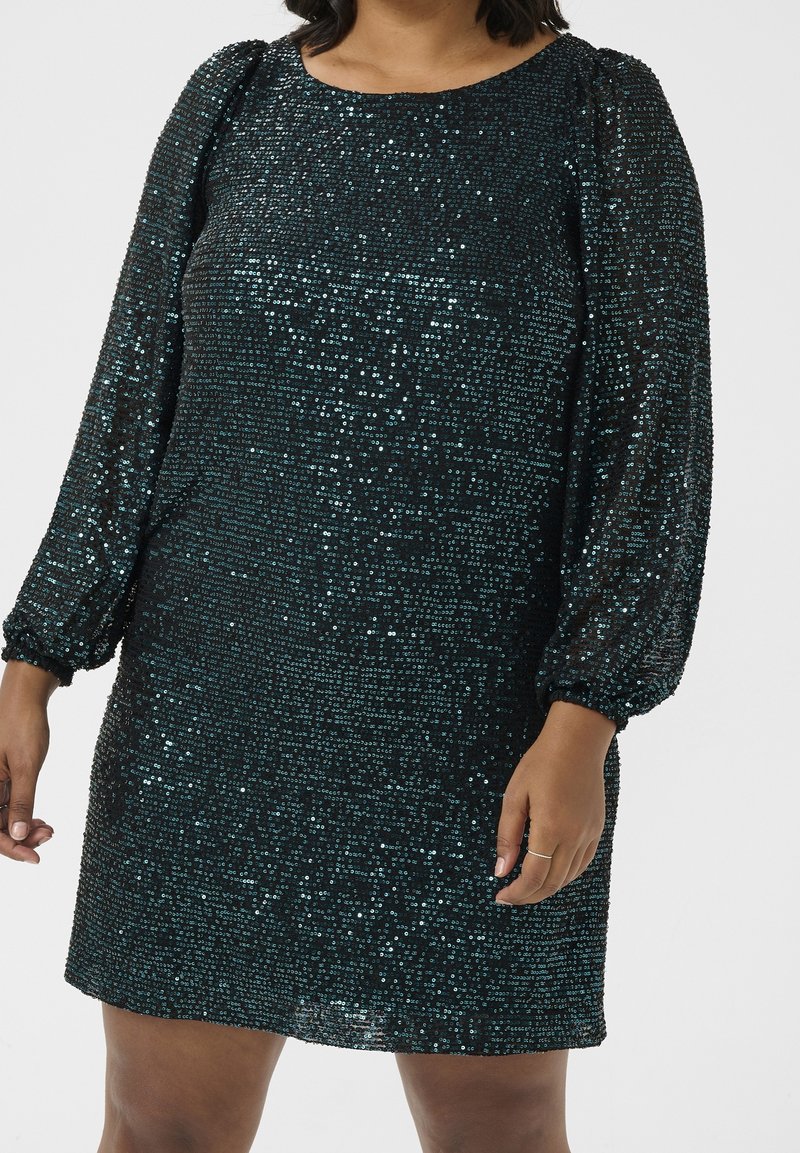 Femme portant une robe noire à manches longues ornée de sequins, avec un col rond et des manches bouffantes, debout devant un fond uni.