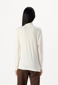 Langermet turtleneck topp i kremfarge med en myk tekstur, som har en avslappet passform og rundet nederkant, sammen med brune bukser.