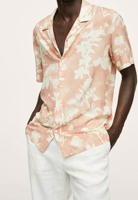 Personne à la peau foncée portant une chemise à manches courtes couleur pêche avec imprimé floral blanc et un pantalon blanc, mains dans les poches, debout devant un fond uni.