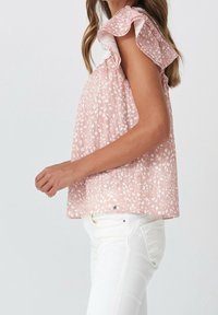 Frau in einer rosa, gemusterten ärmellosen Bluse mit Rüschenschultern und weißen Hosen, von der Seite vor einem schlichten Hintergrund gezeigt.