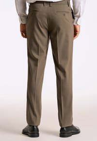 Bruine op maat gemaakte broek met een gestructureerde stof, twee achterzakken met knopen en een rechte pasvorm. Gecombineerd met zwarte schoenen.