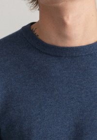 Blauer Strickpullover mit Rundhalsausschnitt. Der Stoff wirkt weich und hat eine leicht strukturierte Oberfläche. Sichtbares Tattoo am Hals.