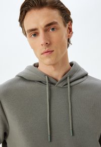 Sudadera de algodón gris con capucha y cordón, que presenta una textura suave y costuras planas y cosidas. Sin logotipos ni patrones visibles.