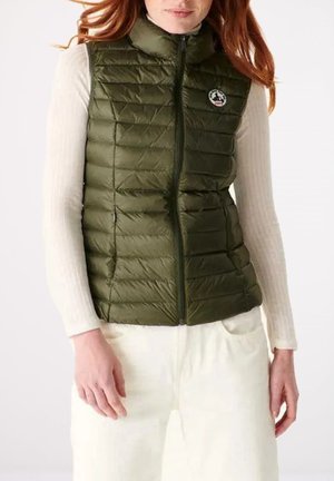 Mujer con un chaleco acolchado sin mangas de color verde oliva sobre una camiseta de manga larga blanca de canalé y pantalones de color claro.