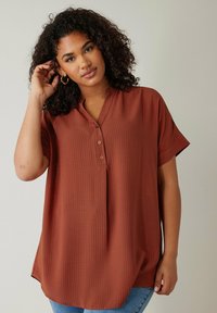 Camisa de botones de color óxido con mangas cortas, fabricada en un tejido texturizado y con un corte relajado. Presenta un cuello y un dobladillo redondeado, combinada con jeans azules.