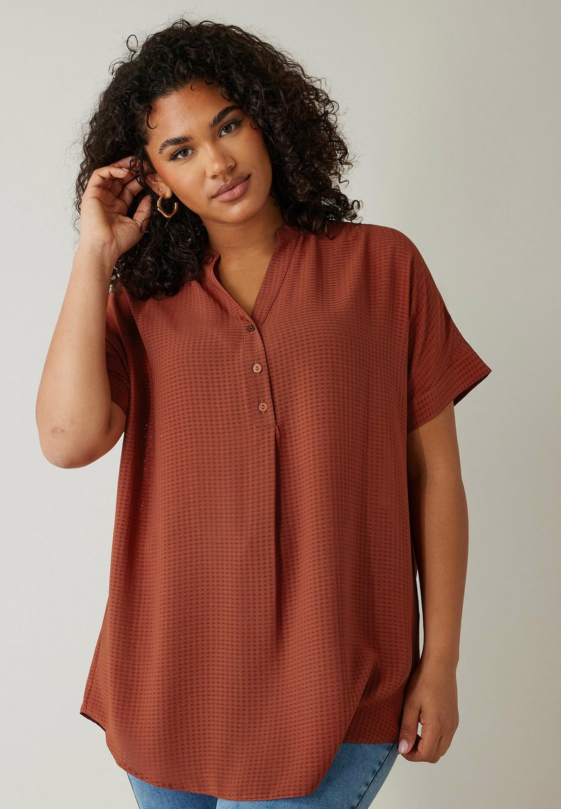 Camisa de botones de color óxido con mangas cortas, fabricada en un tejido texturizado y con un corte relajado. Presenta un cuello y un dobladillo redondeado, combinada con jeans azules.