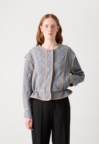 Grijze gewatteerde jas met een textuurpatroon van bladeren, beige afwerking en peplumontwerp. Lange mouwen en een ronde halslijn. Gepaarde met zwarte broek.