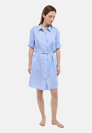 Femme portant une robe chemise bleu clair à boutons, manches retroussées et ceinture nouée à la taille, debout pieds nus devant un fond uni.