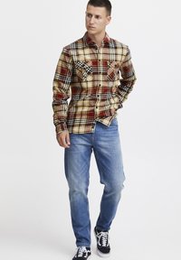 Camisa de franela en cuadros amarillos, rojos y negros con dos bolsillos en el pecho, combinada con jeans azul claro y zapatillas negras.