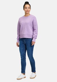 Lavendelfarbenes Strickoberteil mit rundem Halsausschnitt und gerippten Bündchen, kombiniert mit dunkelblauen Jeans und weißen Sneakers mit einer hellbraunen Sohle.