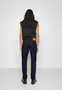 Vaqueros de mezclilla azul oscuro con un corte recto, presentan un parche de cuero en la cinturilla trasera y bolsillos traseros clásicos, combinados con una blusa sin mangas negra.