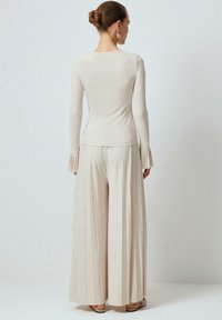 Žena s vlasmi zviazanými do drdola, oblečená v béžovom top s dlhými rukávmi s volánikmi na manžetách a v zodpovedajúcich širokých plisovaných nohaviciach, stojaca chrbtom k nám.